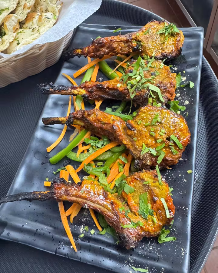 Tandoori lamb chops on a black platter with carrot and green pepper strips, a basket of naan behind them.